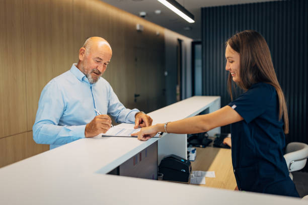 A cheerful nurse at the clinic reception explains to a senior patient how to fill out a form before starting his hospital treatment